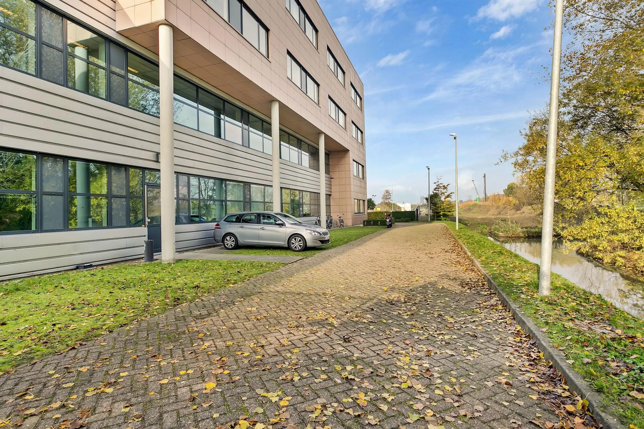 Kantoorgebouw aan de Laan van ‘s-Gravenmade met geparkeerde auto en herfstbladeren op de stoep langs een water.